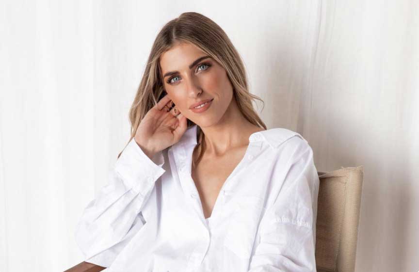 A woman with long hair wearing a white shirt is seated, resting her head on her hand, and smiling softly against a light background, illustrating a relaxed moment ideal for a social media planner | plannthat.com